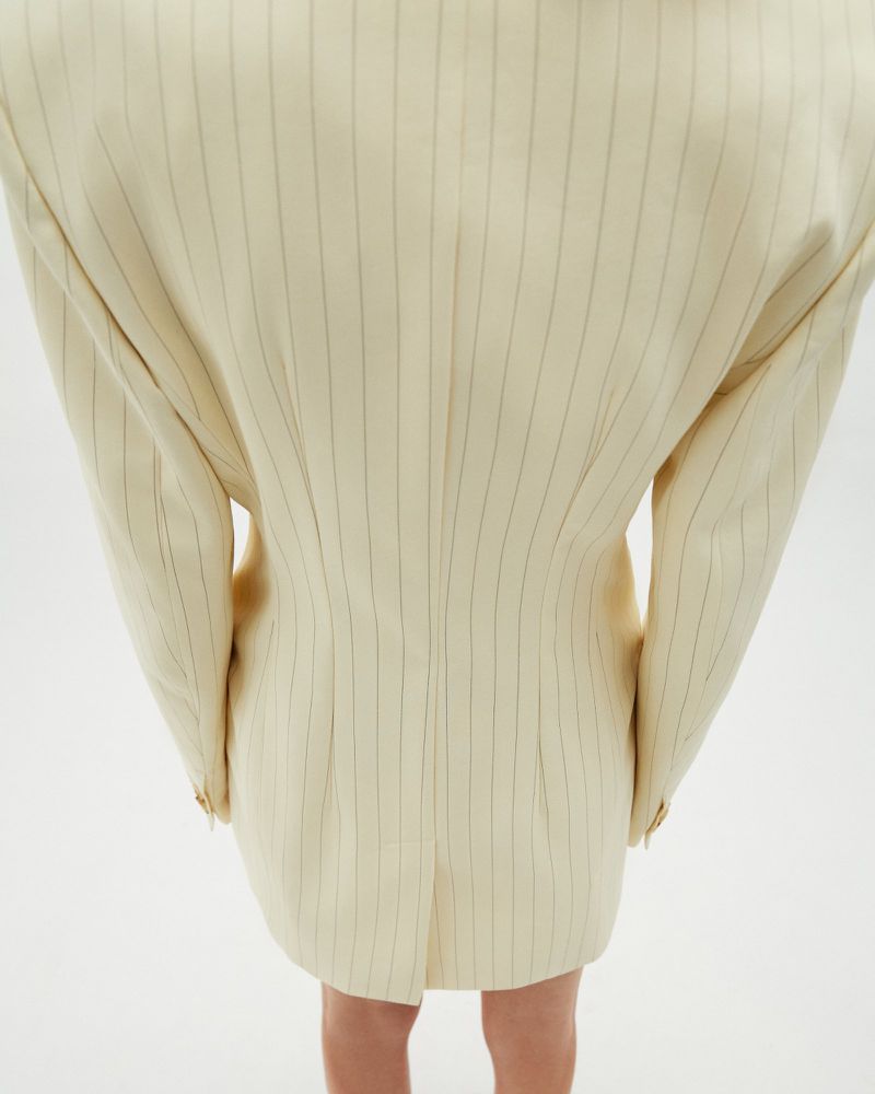 Back view of an adult female wearing a cream-colored, knee-length blazer dress with thin, vertical black stripes against a plain white backdrop.