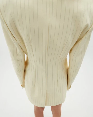 Back view of an adult female wearing a cream-colored, knee-length blazer dress with thin, vertical black stripes against a plain white backdrop.
