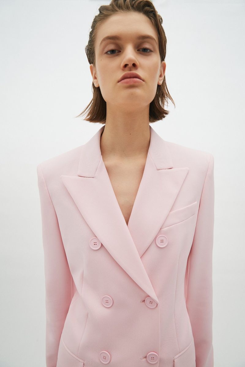 Close-up of a model wearing a double-breasted pink blazer, posing in front of a white background with a neutral facial expression.
