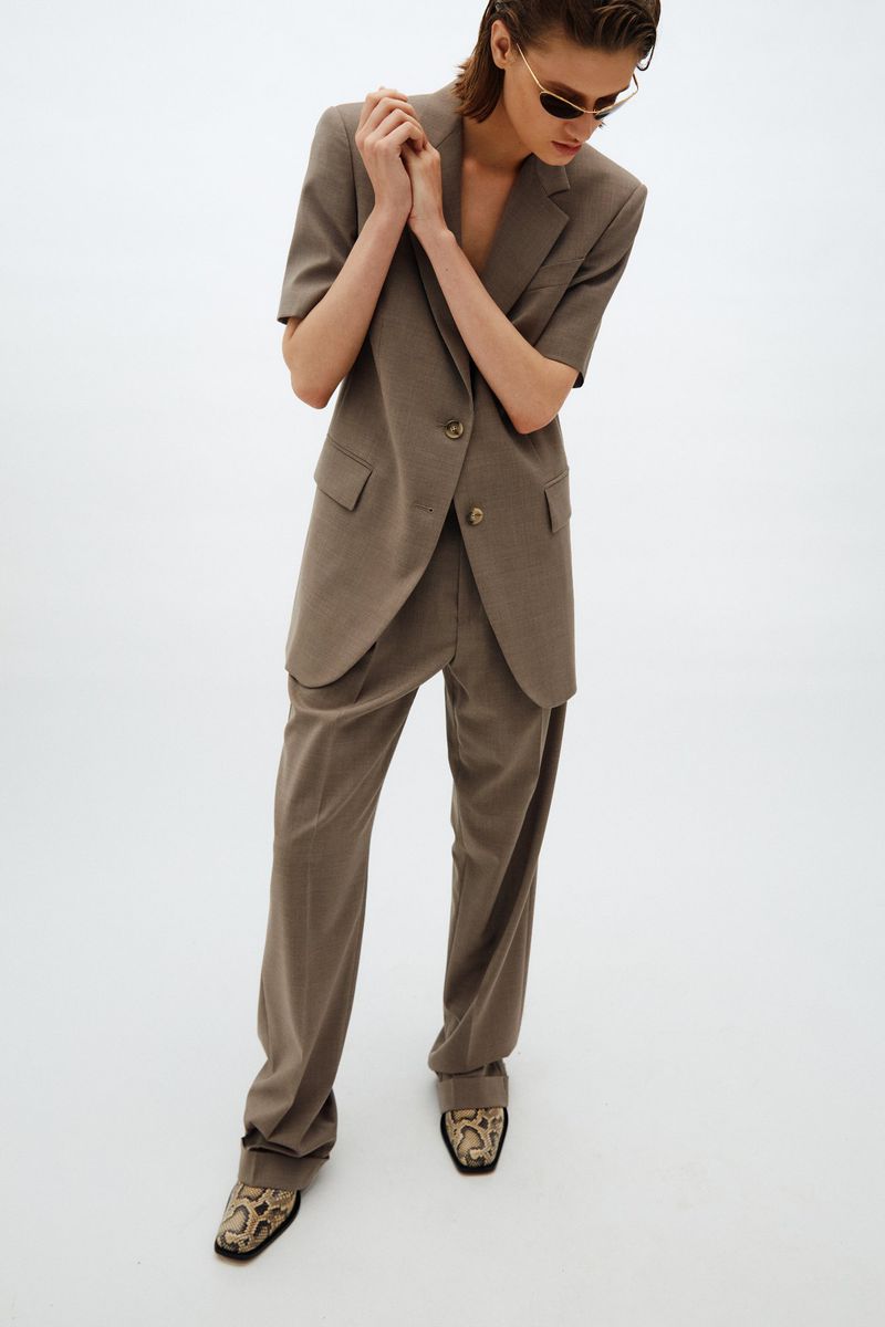 An adult woman stands indoors in a light studio, wearing a brown suit with a short-sleeved jacket, baggy pants, and snake print shoes.