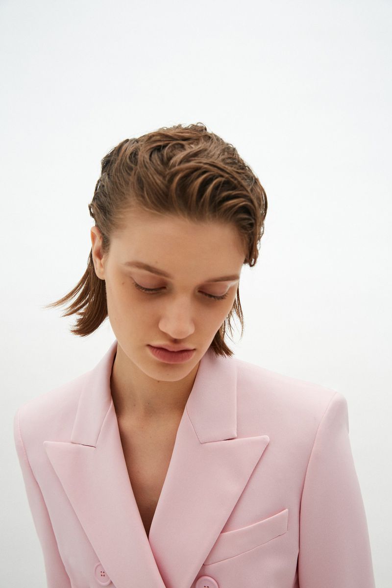 Close-up studio shot of a young adult female model with short wet-look hair wearing a pale pink blazer against a bright white background.