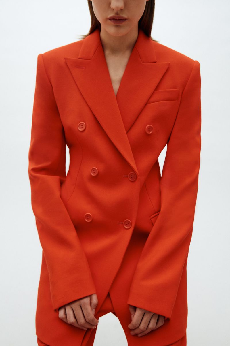 Close-up of a woman in a vibrant orange double-breasted blazer and matching pants, posing in a studio with a clean, white backdrop.