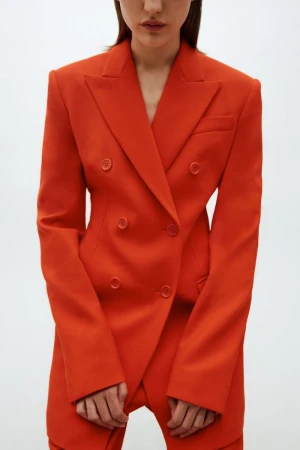 Close-up of a woman in a vibrant orange double-breasted blazer and matching pants, posing in a studio with a clean, white backdrop.