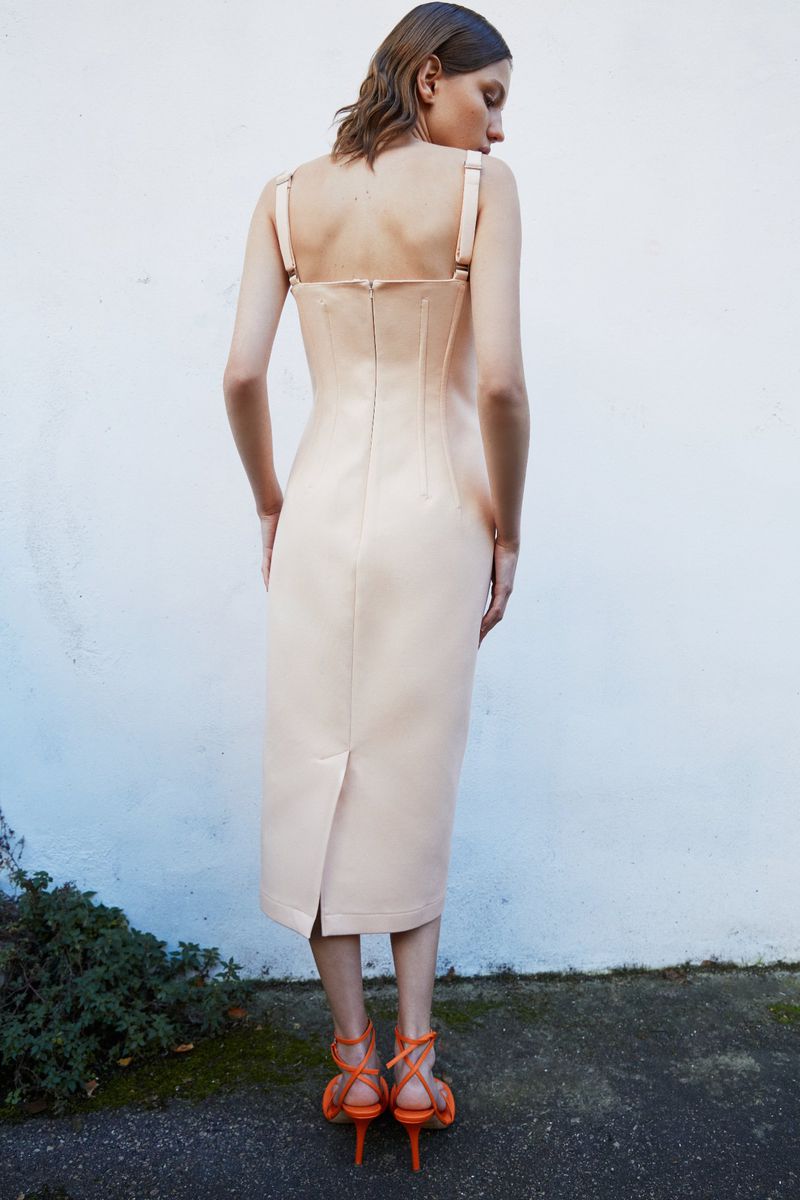 Woman in light pink midi dress and orange strappy heels stands against a textured white wall with green foliage at the base.