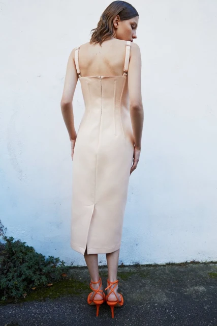 Woman in light pink midi dress and orange strappy heels stands against a textured white wall with green foliage at the base.