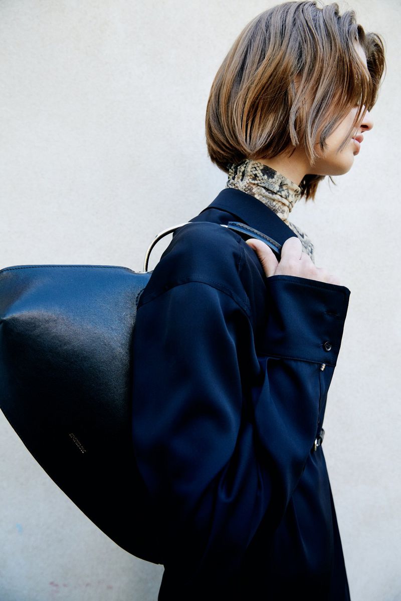 Brunette woman in black jacket and patterned top carrying a black tote bag stands against a white wall in an outdoor setting.