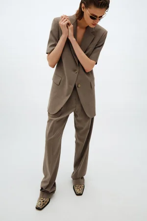 Model wearing a brown short-sleeve blazer and matching trousers with snakeskin shoes against a white backdrop