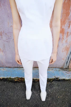 Model standing outdoors wearing a white sleeveless top and matching white trousers against a weathered wall