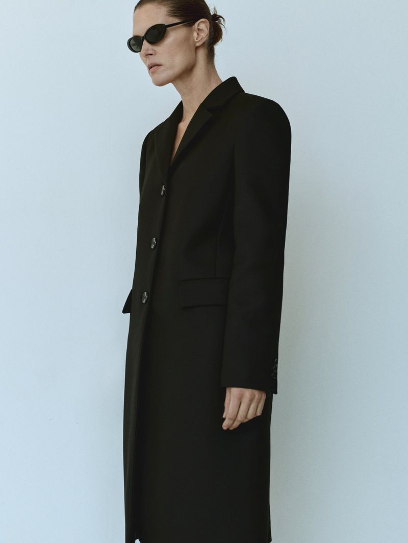 Adult female posing in long black overcoat and oval sunglasses against a plain off-white background, portrait fashion shot.