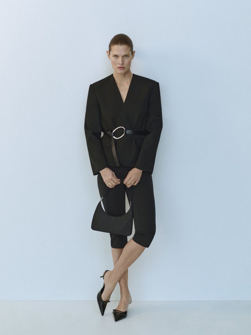 Elegant woman poses in a black pantsuit with a silver belt buckle, holding a black purse, wearing black heels against a plain backdrop.