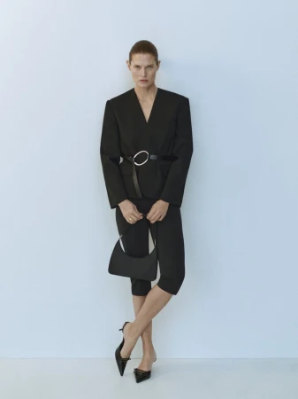 Elegant woman poses in a black pantsuit with a silver belt buckle, holding a black purse, wearing black heels against a plain backdrop.