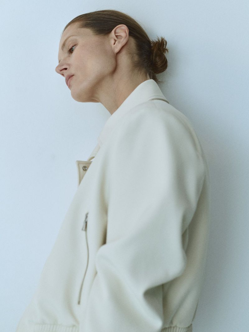 Woman in a cream-colored bomber jacket posing against a plain white background. Studio shot, fashion-forward, minimalist aesthetic.