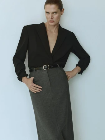 Fashion shoot of a slim adult woman in a black jacket, dark grey skirt, and belt against a neutral wall backdrop in a photo studio.