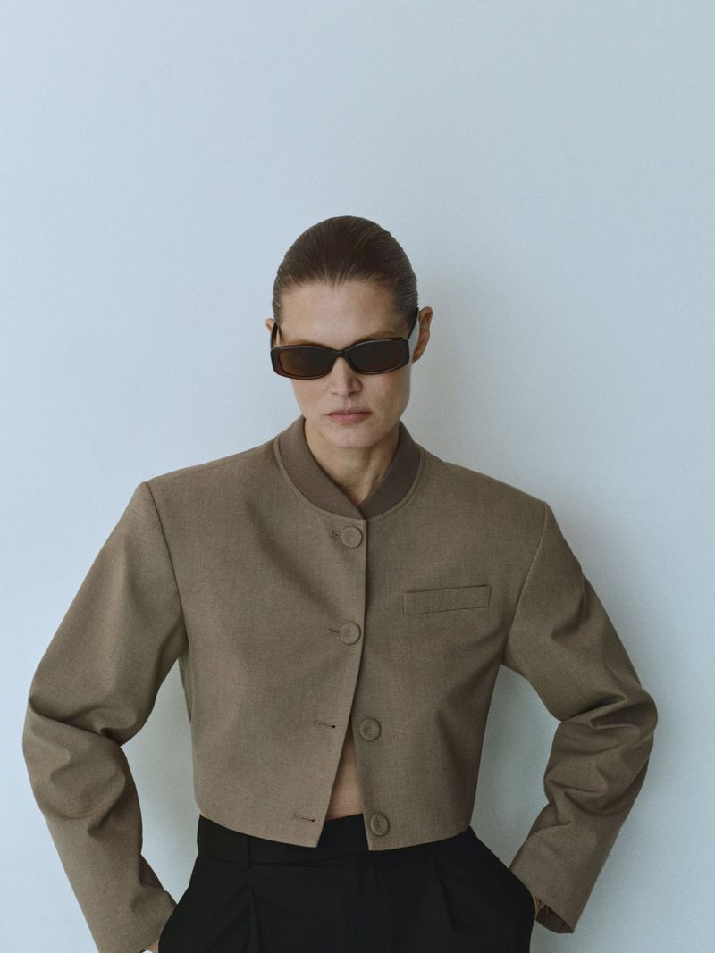 A woman wears a brown cropped jacket and sunglasses against a white background, presenting a modern, minimalist fashion aesthetic.