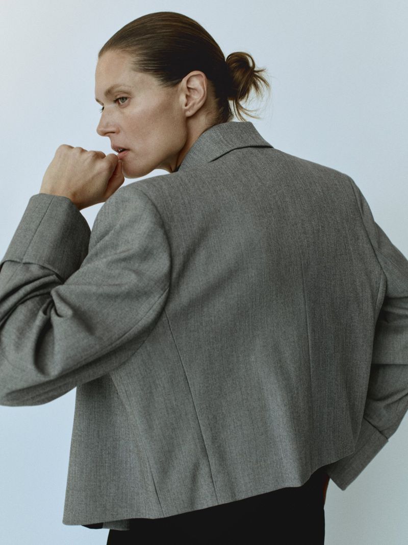 Adult woman in a gray blazer in an indoor setting, looking to the side with her hand near her mouth, side shot, fashion concept.