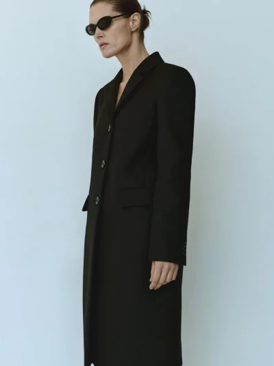 Model Malgosia Bela wearing a long black coat and dark sunglasses against a pale neutral background