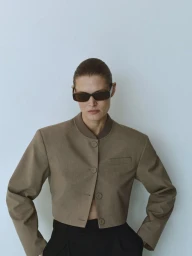 Model wearing dark sunglasses and a brown buttoned jacket against a pale grey background