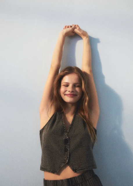 Portrait of a young woman with auburn hair, wearing a waistcoat, stretching her arms overhead against a pale blue wall in studio lighting.