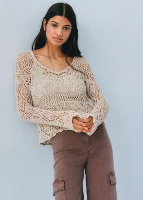 Indoor studio portrait of a young adult woman wearing a tan knit sweater and brown pants against a light blue background.