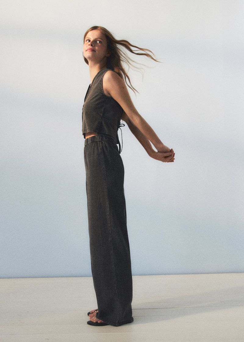 A young woman models sustainable fashion in a grey crop top and wide-leg pants, arms behind her back, hair blowing in the breeze, studio shot.