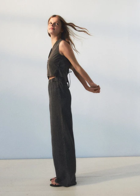 A young woman models sustainable fashion in a grey crop top and wide-leg pants, arms behind her back, hair blowing in the breeze, studio shot.