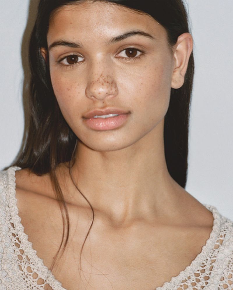 Close-up portrait of a woman with brown hair and freckles, wearing a cream-colored lace top, against a light background in soft lighting.