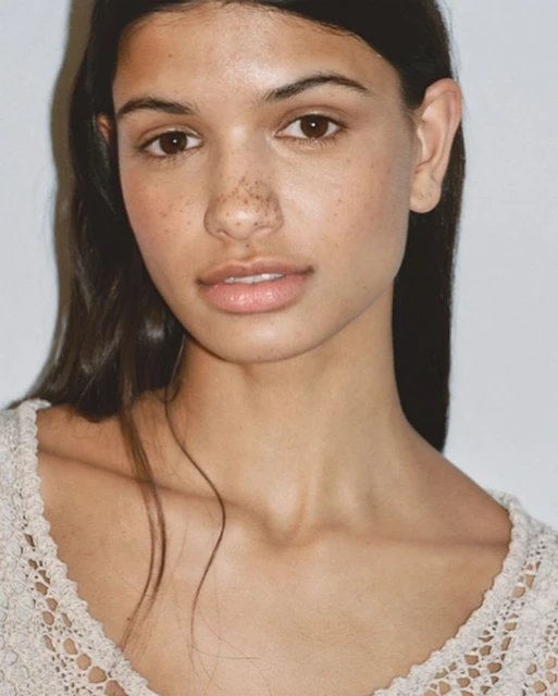 Close-up portrait of a woman with brown hair and freckles, wearing a cream-colored lace top, against a light background in soft lighting.