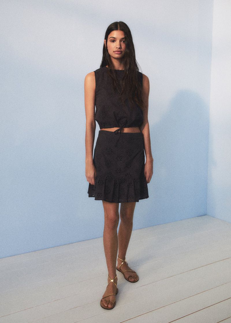 Brunette woman poses in a sleeveless crop top and mini skirt with ruffles in a studio with a blue and white wall.