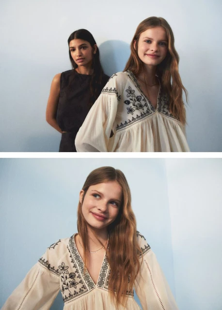 Two women pose indoors against a soft blue wall, one wearing an embroidered peasant blouse and the other a dark sleeveless top and pants.