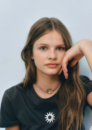 Close-up portrait of a young woman with long brown hair wearing a black t-shirt with a white sun graphic and a necklace.