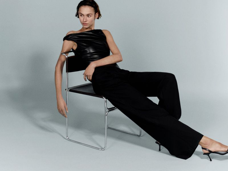Elegant woman in black top and wide-leg pants poses while seated on a black and chrome chair in a minimalist studio setting.