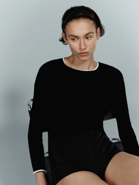 A woman with short dark hair sits in a chair wearing a long-sleeved black top with white trim and black shorts against a neutral background.