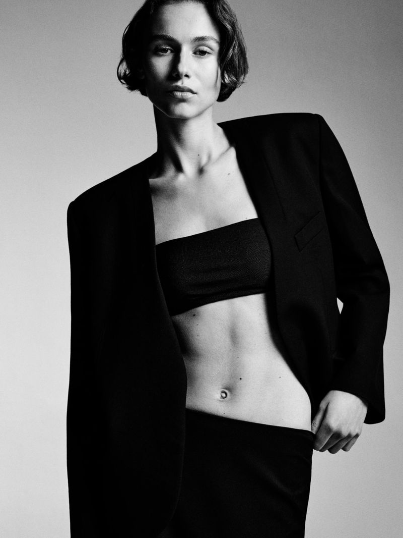 Black and white portrait of a young woman in a black blazer and tube top, posing against a plain backdrop with studio lighting.