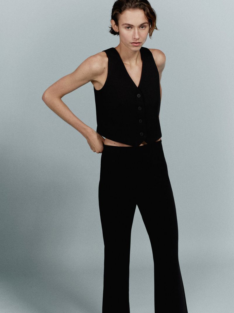 Fashionable young adult woman poses in a sleeveless black vest and matching flared black pants on a neutral grey background.