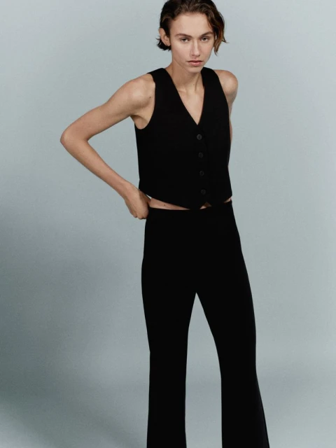 Fashionable young adult woman poses in a sleeveless black vest and matching flared black pants on a neutral grey background.