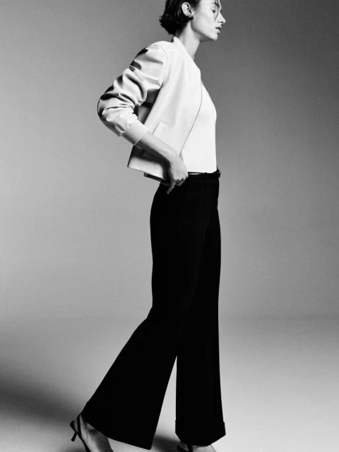 Monochrome full shot of an adult female fashion model in a light jacket and dark pants, posing in a photo studio with side lighting.