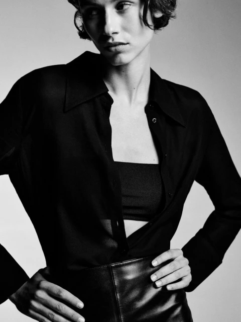 Black and white portrait of a stylish adult woman in an unbuttoned black blouse and skirt, hands on hips, set against a light backdrop.