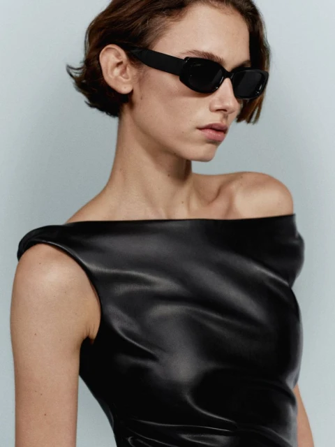 Close-up studio shot of a woman with short hair wearing black sunglasses and a draped black faux leather top on a light blue background.