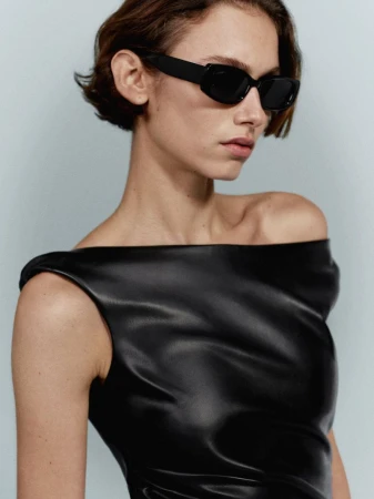 Close-up studio shot of a woman with short hair wearing black sunglasses and a draped black faux leather top on a light blue background.