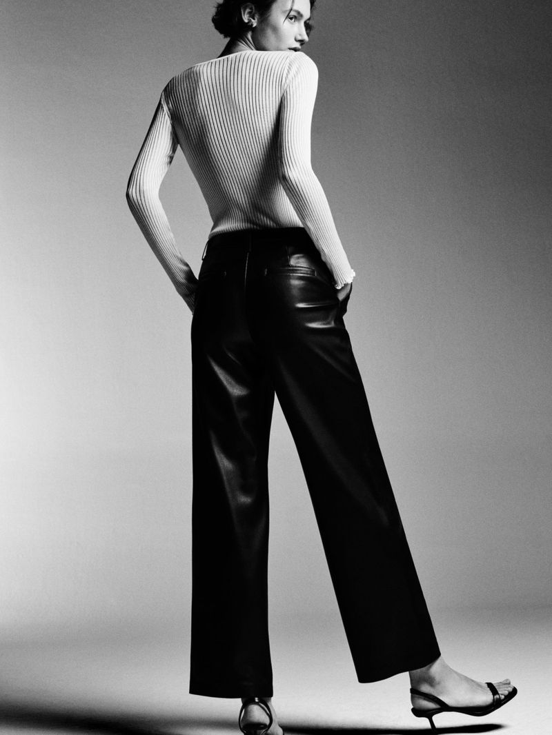 Studio shot of an adult woman posing in a white ribbed top and black leather pants with high heels, shot in black and white.