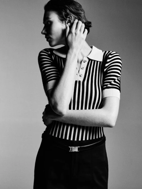 Monochrome portrait of a poised adult woman with short hair wearing a striped top and dark pants against a soft grey background.