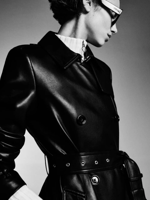 Monochrome fashion portrait of a woman in a black leather trench coat and white-rimmed glasses posing against a neutral gray backdrop.