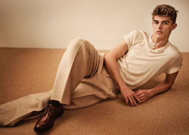 Blonde male model lies on brown carpet in cream shirt and tan pants, posing with a serious expression in indoor photo studio shot.