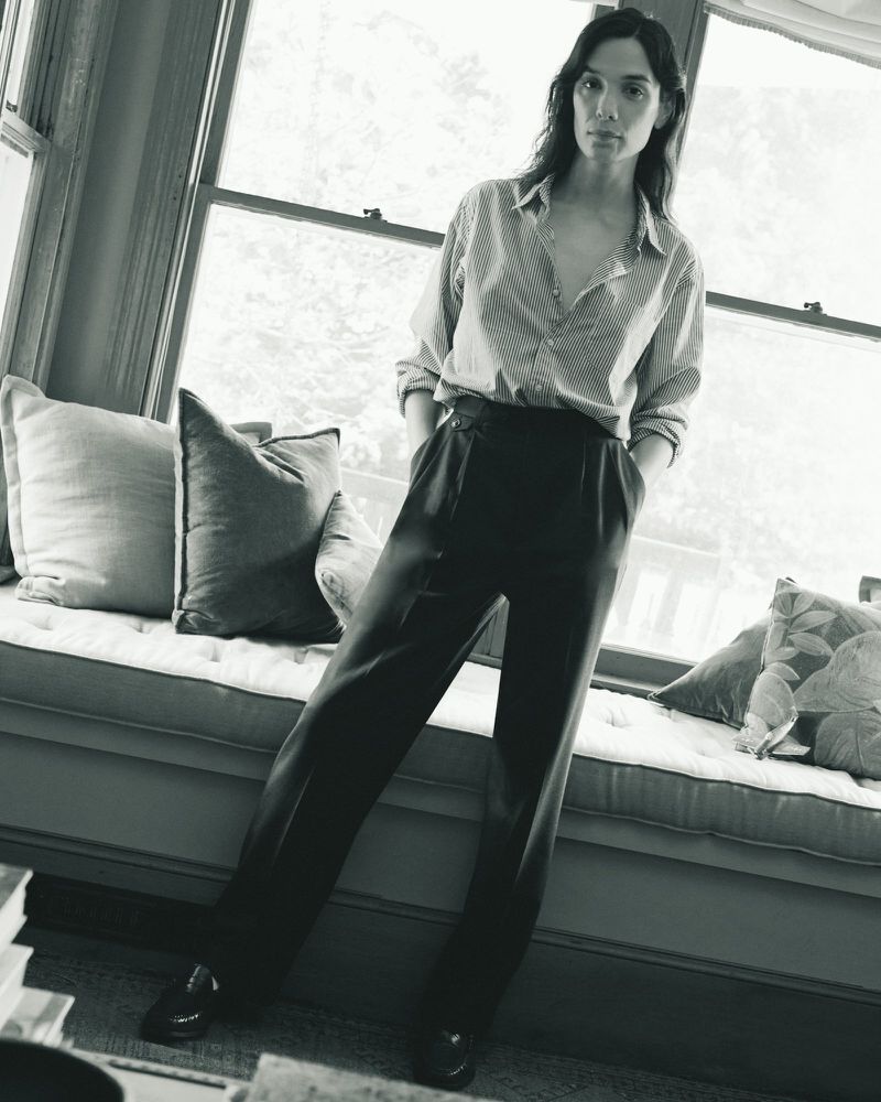 Monochrome portrait of a woman in a striped shirt and dark pants, casually posed by a window with soft, natural light.