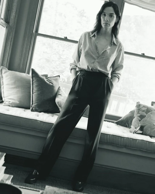 Monochrome portrait of a woman in a striped shirt and dark pants, casually posed by a window with soft, natural light.
