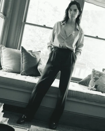 Monochrome portrait of a woman in a striped shirt and dark pants, casually posed by a window with soft, natural light.