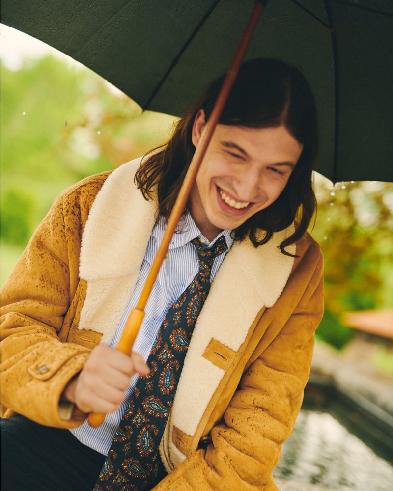 A young adult man with long hair smiles under an umbrella. He wears a shearling-lined coat, striped shirt, and patterned tie on a rainy day.