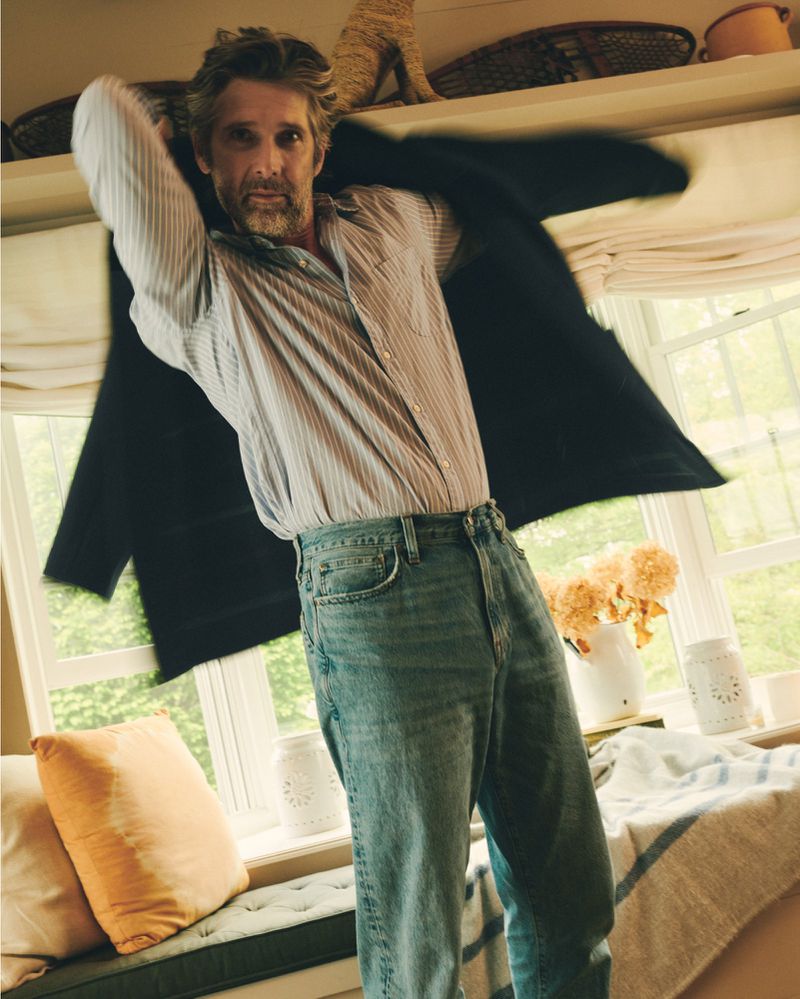 Middle-aged man wearing a striped shirt and jeans puts on a black coat indoors with natural light from a window.