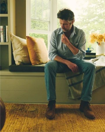 Adult man sitting on a bench in a living room, wearing a collared shirt and jeans, with neutral colors and warm indoor lighting.