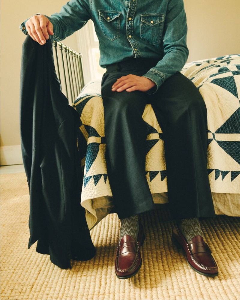 Man sitting on a bed wearing a denim shirt, holding a suit jacket, displaying brown leather loafers and patterned socks in a room interior.